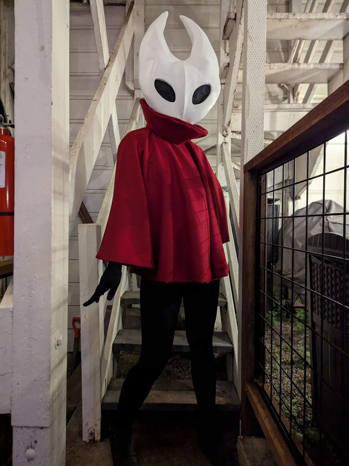 A person in a white Hornet mask with two tall horns, a red cloak with a high collar, black bodysuit and black boots, posing on a wooden staircase