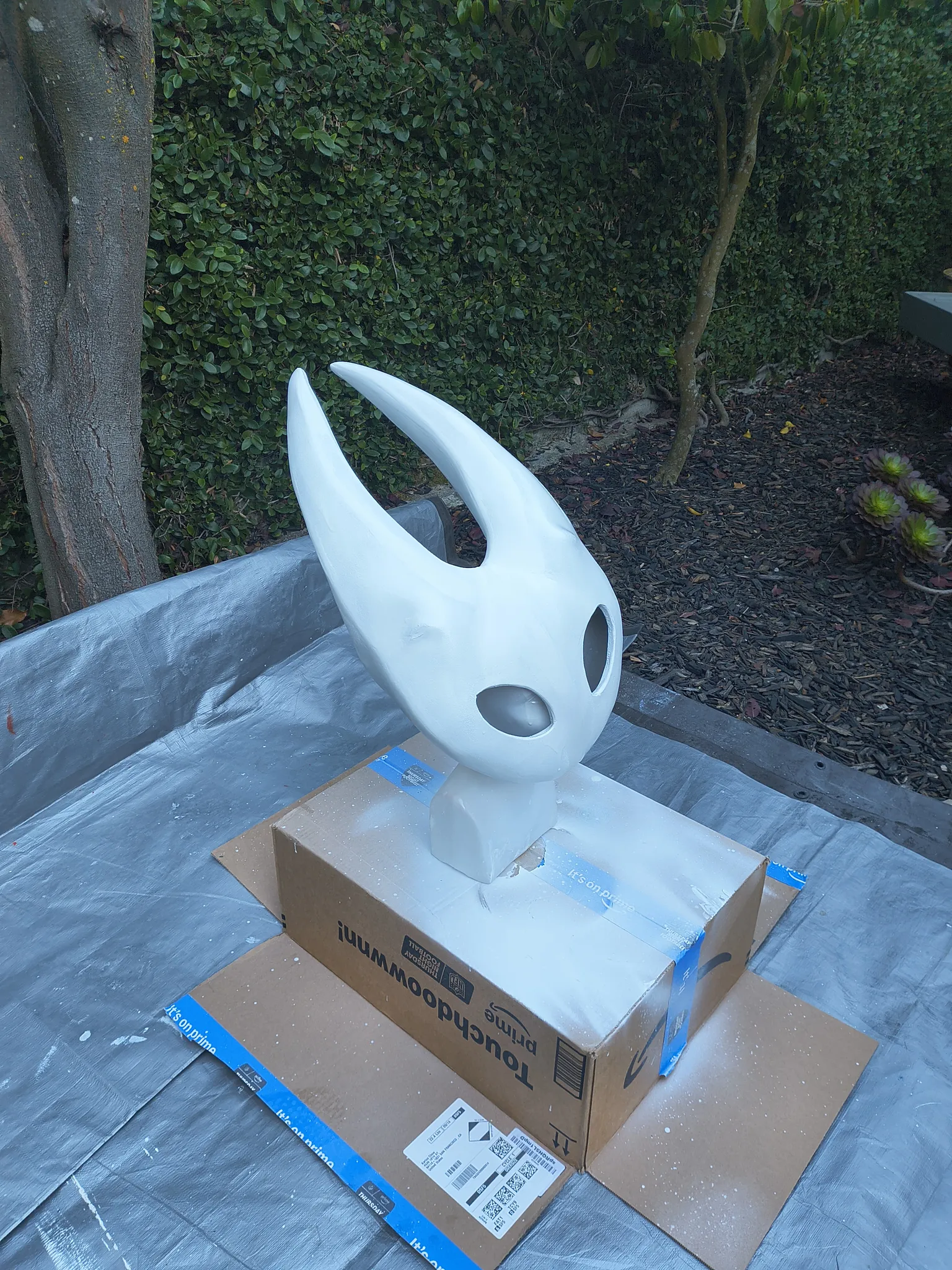 The mask painted white, drying on a cardboard box on a tarp in the garden