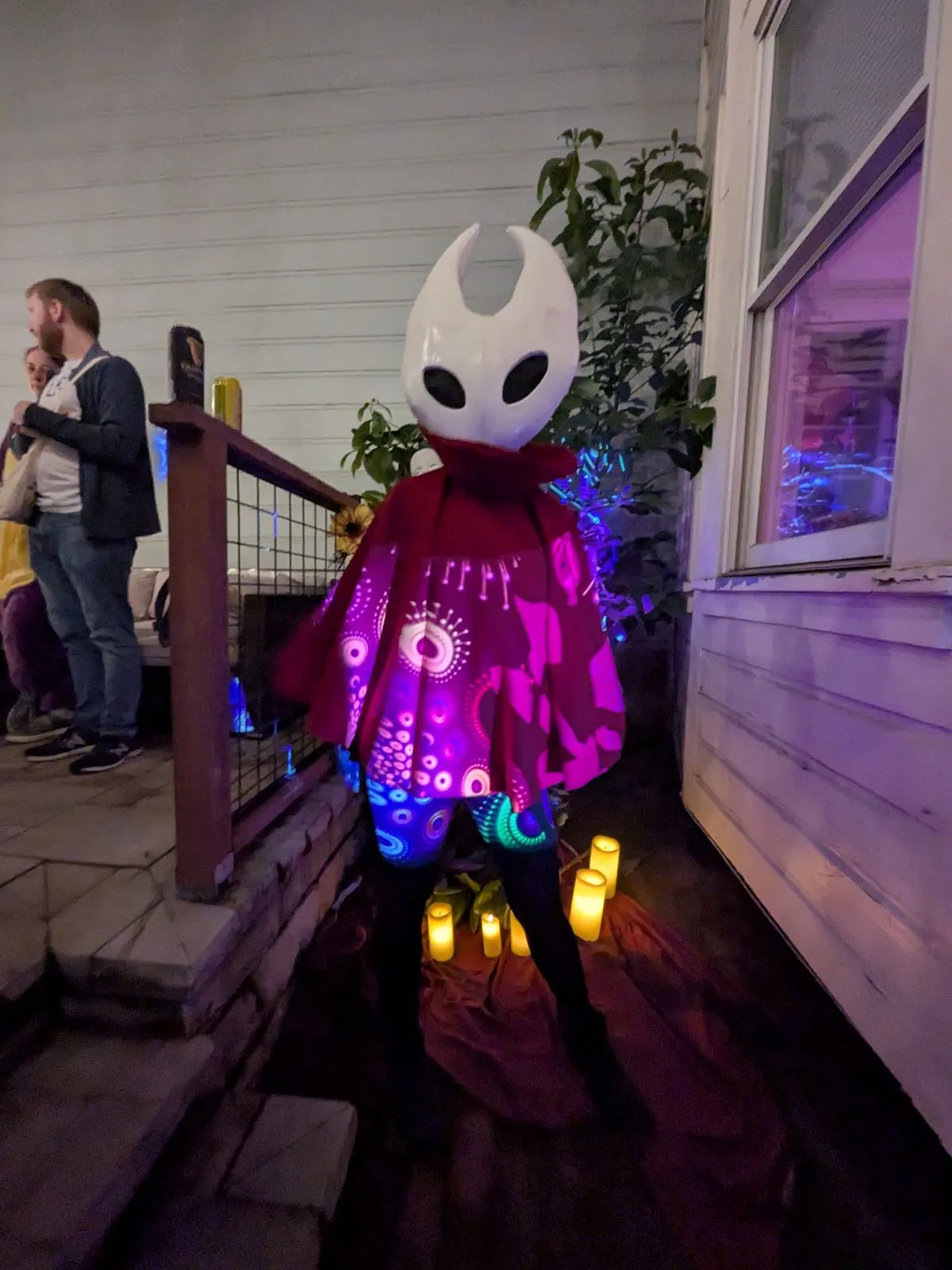 Hornet at a party at night, the red cloak lit purple by string lights, with other guests behind
