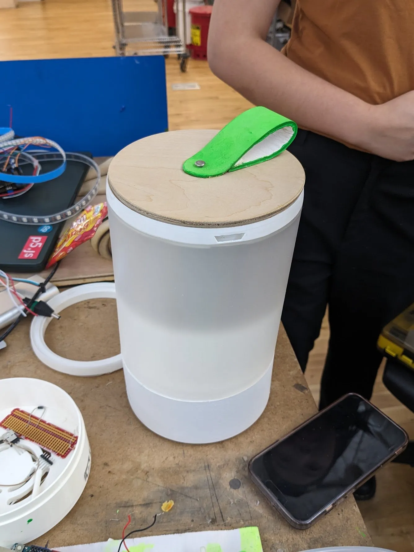 The nearly finished diffuser standing assembled on a desk: white cylindrical shell, wooden lid, and a bright green leather pull tab, with the metal bucket parts beside it.