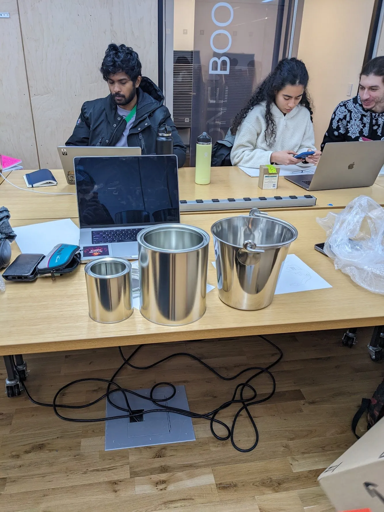 Three metal containers of descending size lined up on a classroom table, with classmates working at laptops in the background.
