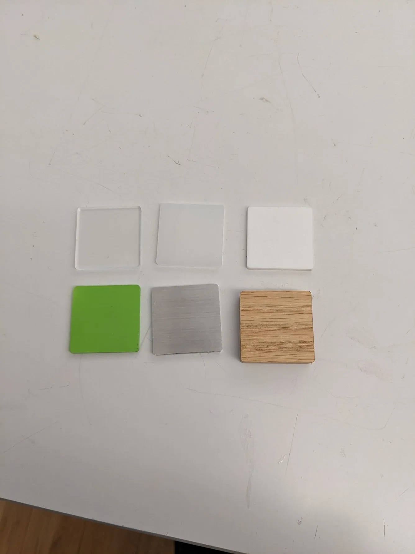 Six color swatch squares laid out on a white surface: three whites and greys on top, with green, grey, and a wood sample below them for matching.