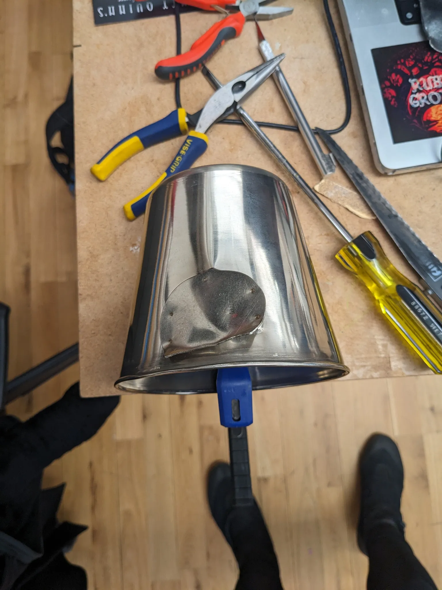 The metal pail on the workbench floor surrounded by pliers and screwdrivers, viewed from above with the maker's boots in frame.