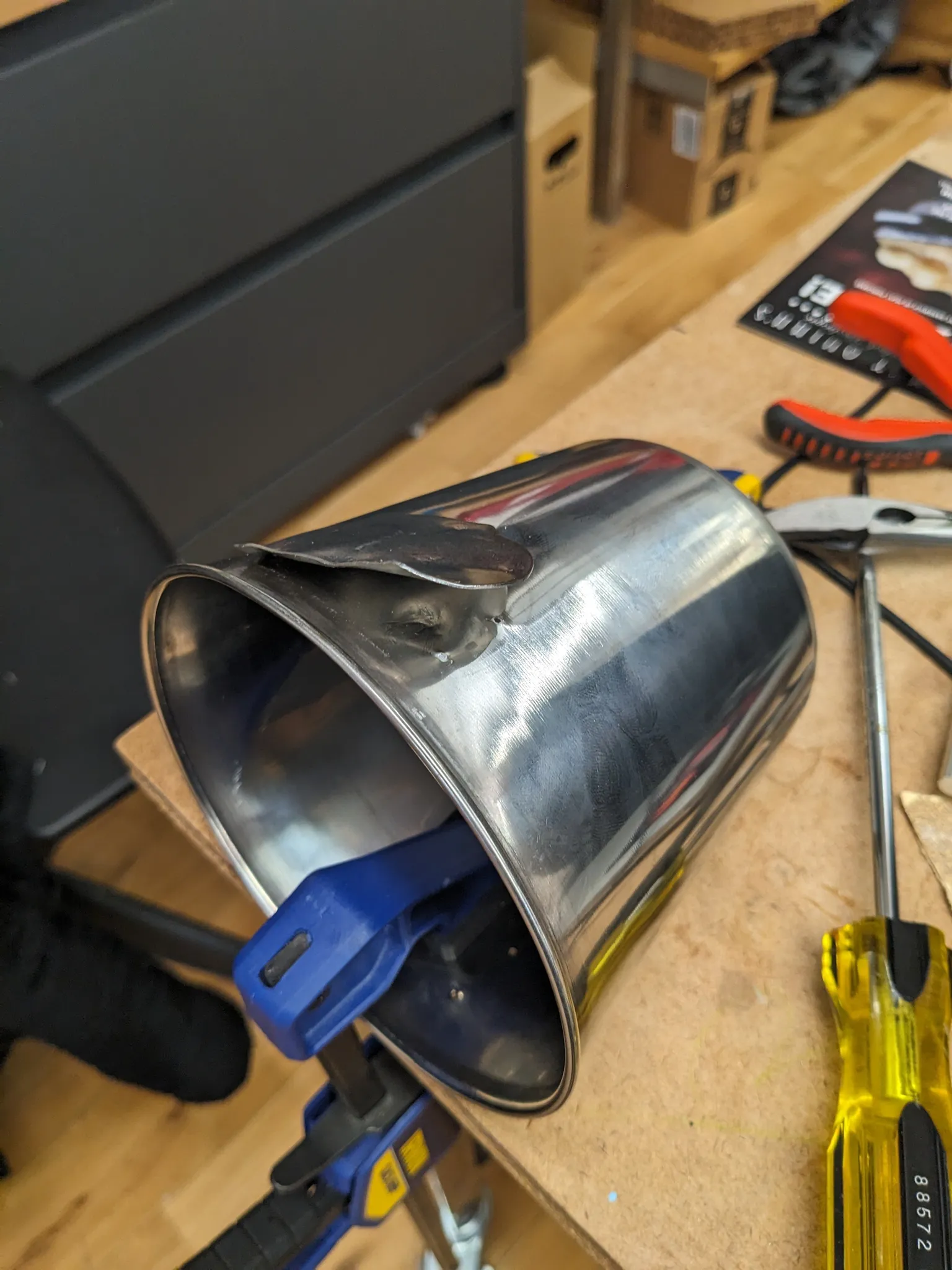 The pail clamped on its side to the bench with a blue clamp while a screwdriver is levered under a welded decoration.
