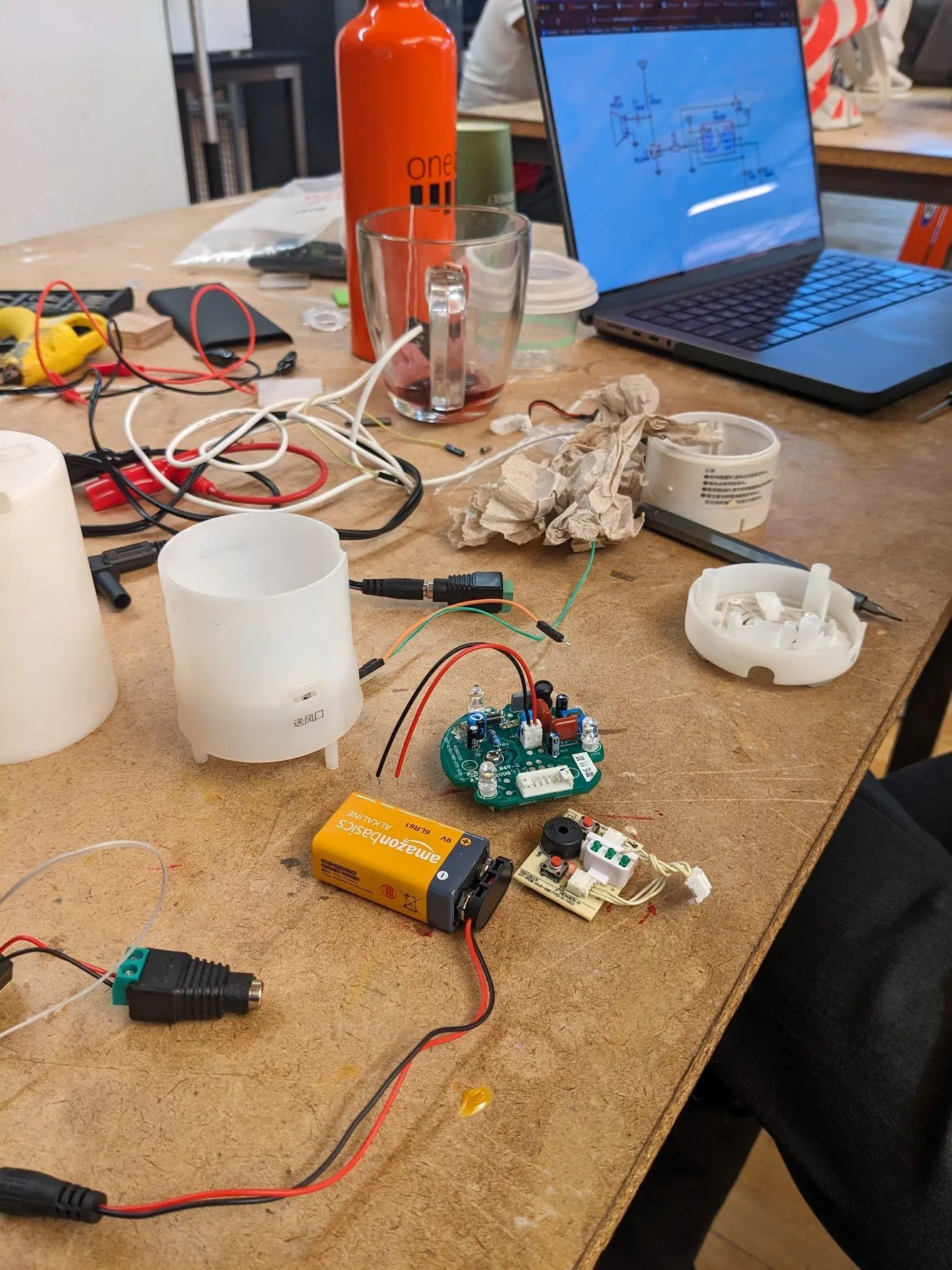 A workbench covered in the disassembled parts of the MUJI diffuser: the white outer shell, a capacitor, two separate circuit boards, and lengths of stripped wire, with a laptop in the background.