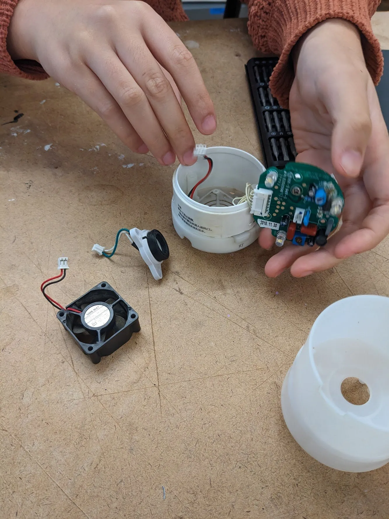 Two hands holding the opened white plastic housing of a MUJI aromatic diffuser, with a small black fan and a green circuit board pulled out beside it on a workbench.