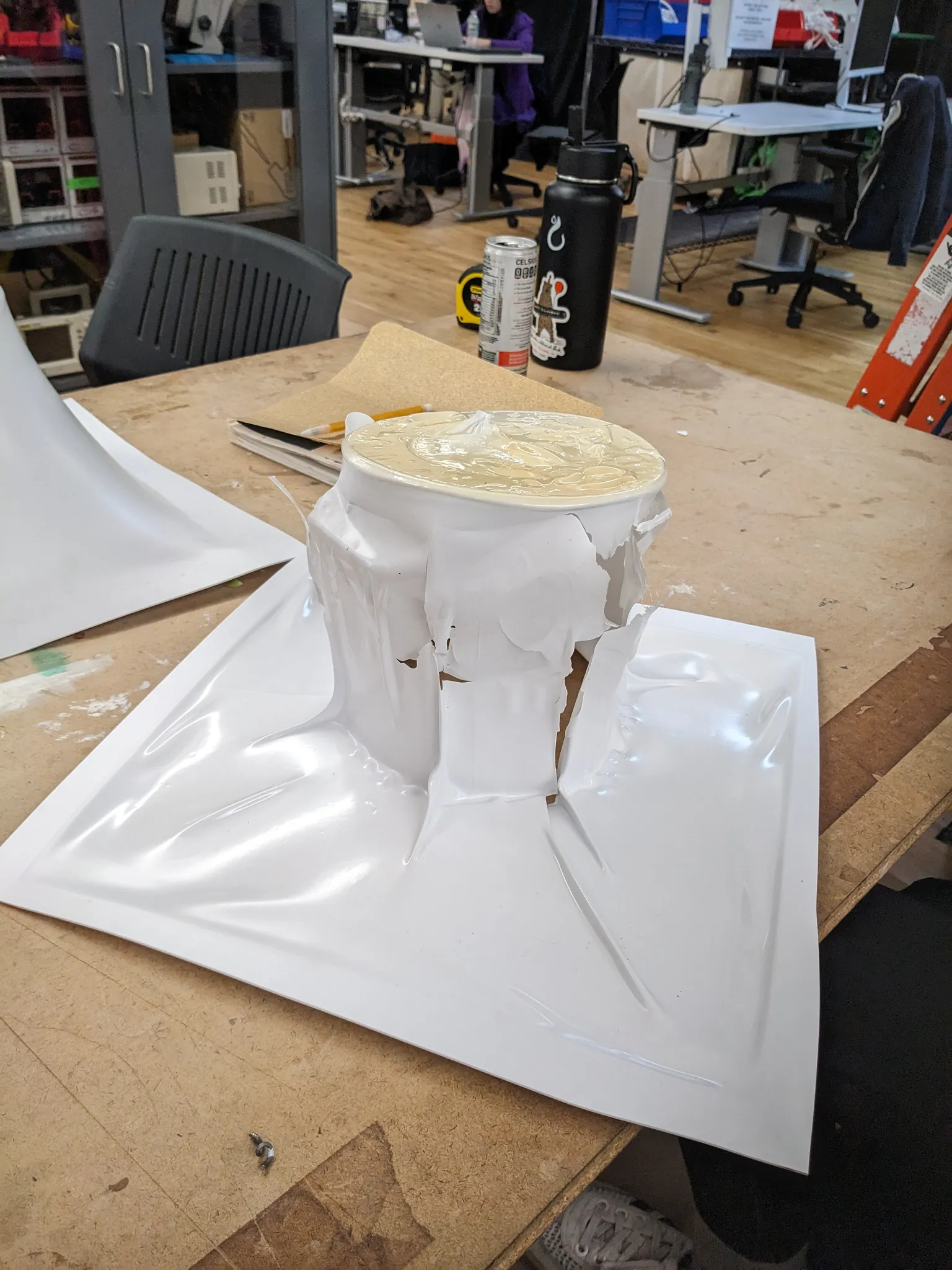 A vacuum formed white plastic shell that has collapsed and crumpled around the can it was formed over, sitting on a sheet of plastic on the workbench.