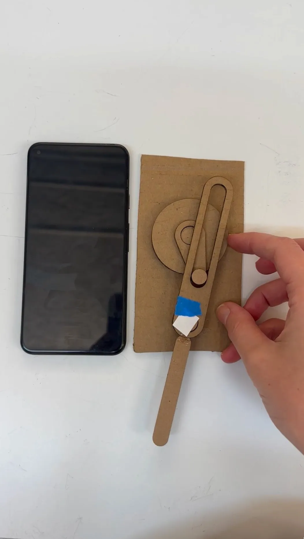 A hand holding a flat cardboard mockup with a long slot cut for the pendulum arm, next to a phone for scale.