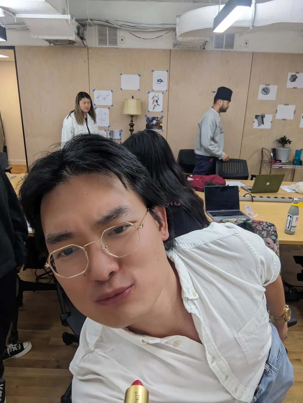 A person in the studio holding the lipstick tube up, classmates working at desks in the background.