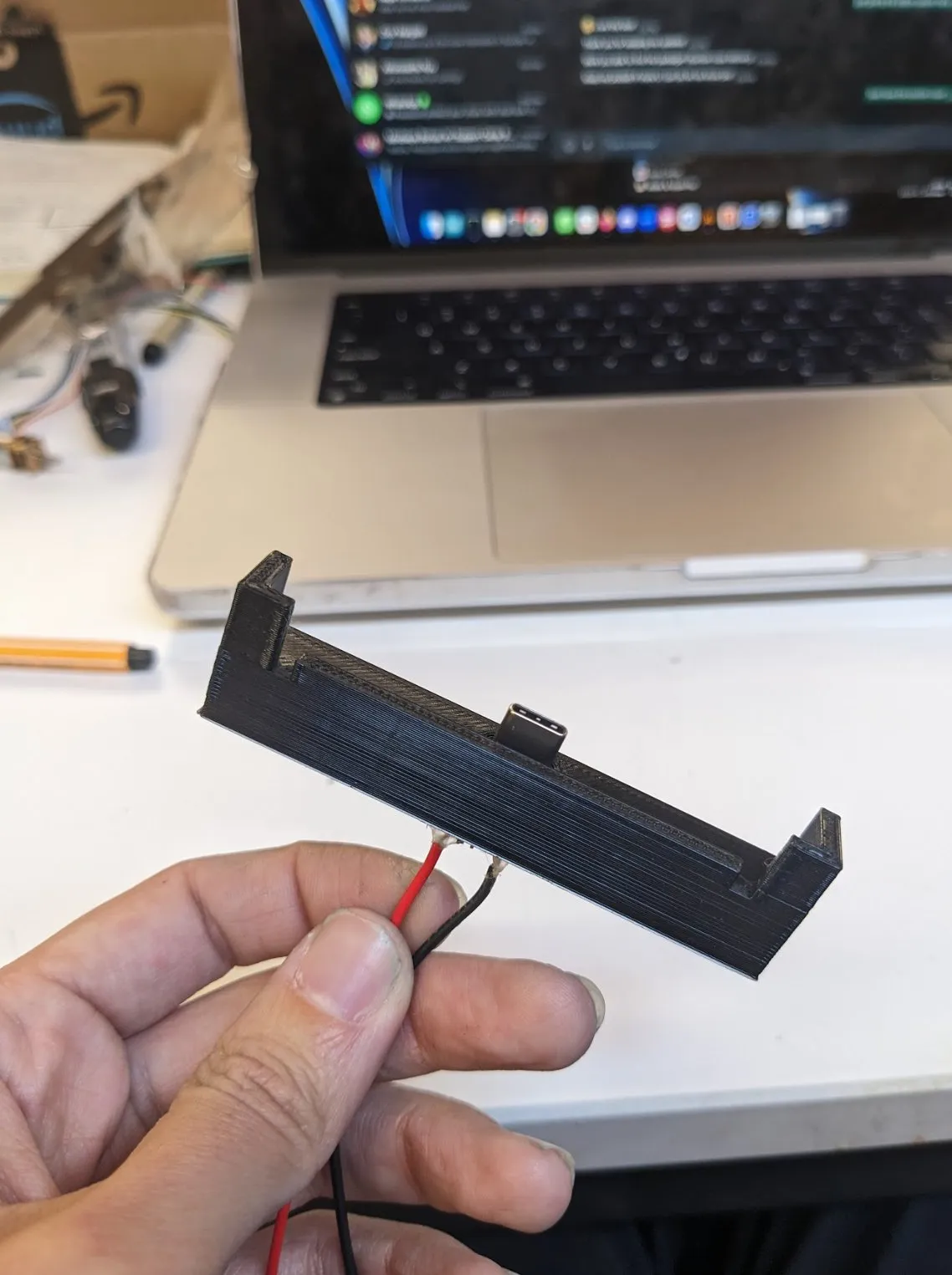 A hand holding the printed black frame piece with thin wires threaded through it, laptop on the desk behind.