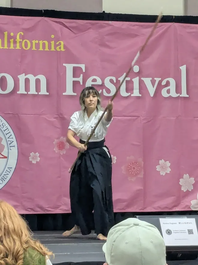 A single practitioner in keikogi and hakama mid-demonstration on the Cherry Blossom Festival stage, naginata raised diagonally overhead, the pink festival banner filling the frame behind them.