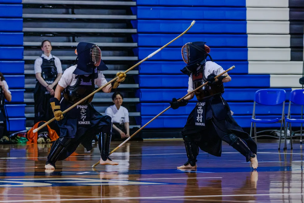 Two practitioners in full bogu facing off on a competition court, one dropped low and one stepping through, their naginata crossing in a wide X between them.